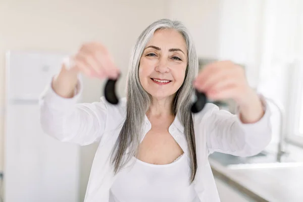 Portrait of pretty smiling grandmother with long gray hair, wearing white shirt, holding black eye patches, showing it to camera, posing over light kitchen background