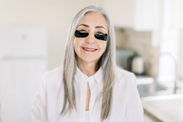 Facial mask, eye patches and cosmetology concept. Attractive elderly woman with beautiful long gray hair, standing with black eye patches at home and enjoying her spa procedures — Stock Photo, Image