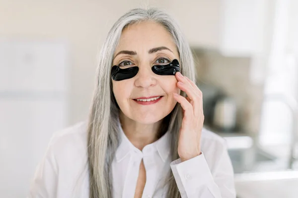 Close up beauty portrait of smiling pretty senior gray haired woman, applying under eye mask, black patches on her face with hands, posing to camera on the background of home interior — Stock Photo, Image