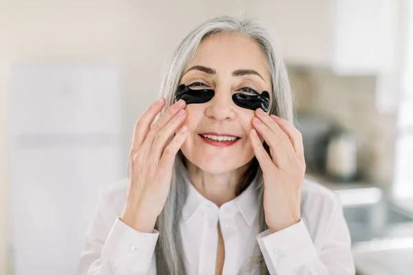 Close up beauty portrait of smiling pretty senior gray haired woman, applying under eye mask, black patches on her face with hands, posing to camera on the background of home interior — Stock Photo, Image