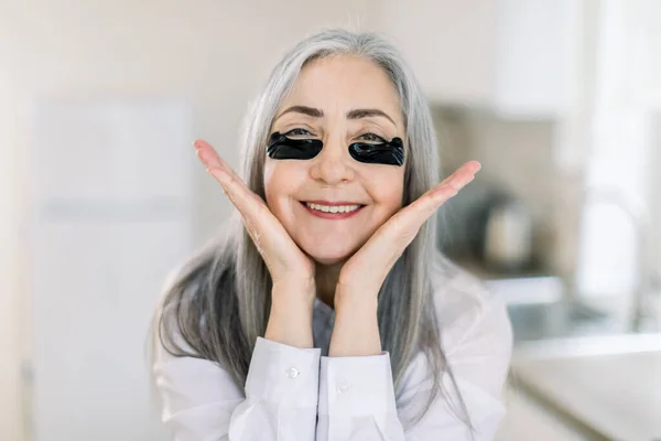 Beauty, antiaging skin care, and senior people concept. Close up portrait of smiling senior woman with black hydrogel under-eye patches touching her face over home interior background — Stock Photo, Image