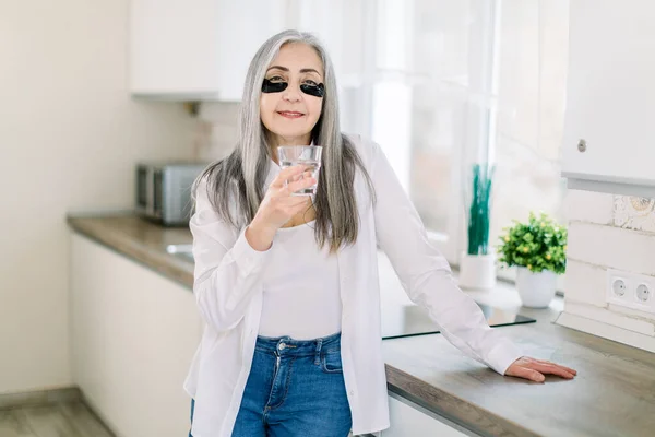 Age, health and people concept. Skin care and beauty routine. Healthy lifestyle and nutrition. Happy senior woman with glass of water and black eye patches posing to camera in kitchen at home — Stock Photo, Image