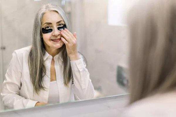 Under eye masks for puffiness, wrinkles, dark circles. Eye patches concept. Reflection in the mirror of smiling senior gray haired woman in white shirt, applying black eye patches in bathroom