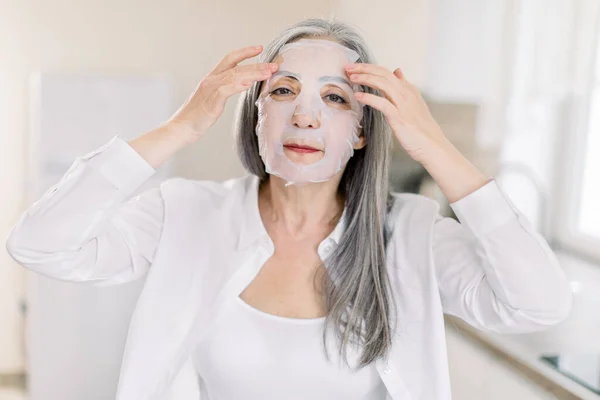 Pretty elderly gray haired woman puts cloth nourishing mask on her face skin for rejuvenating and eliminating defects, while standing on the background of light home interior — Stock Photo, Image