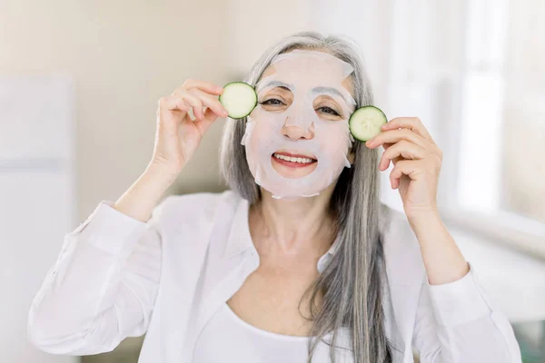 Cuidados com a pele do corpo e do rosto, tratamento anti envelhecimento. Close up tiro horizontal de mulher muito sênior com máscara facial pano, segurando fatias de pepino, enquanto em pé sobre o fundo interior de casa — Fotografia de Stock