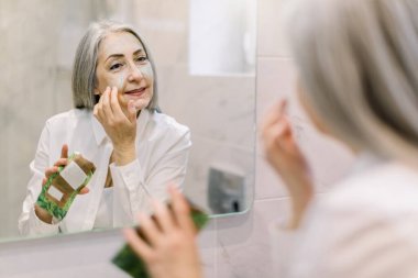 Beyaz gömlekli, gülümseyen, banyo aynasına bakarak cilt maskesi takan güzel bir emekli kadın. Sabah cilt bakımı güzellik rutini, kırışıklık önleyici spa tedavisi konsepti