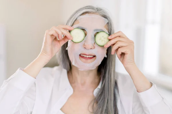 Smiling gray haired senior woman at home, with facial sheet anti-aging mask on face skin, holding fresh cucumber slices, covering her eyes. Beauty and skin care procedures — Stock Photo, Image