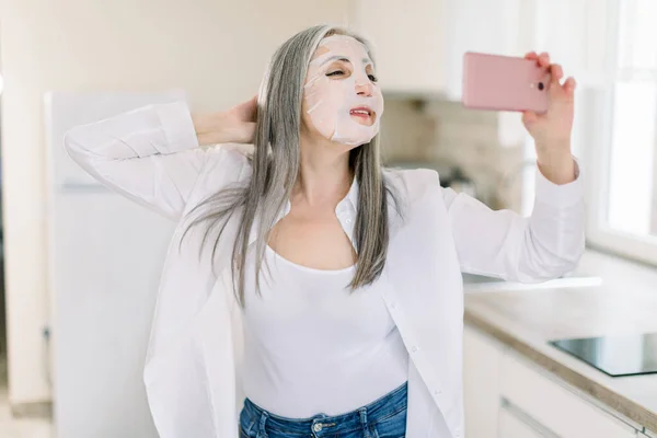 Wanita tua yang menarik dalam kemeja putih dan celana jeans denim, dengan lembar masker wajah di wajahnya, menggunakan smartphone-nya untuk panggilan video atau membuat foto selfie di dapur rumah cahaya modern — Stok Foto