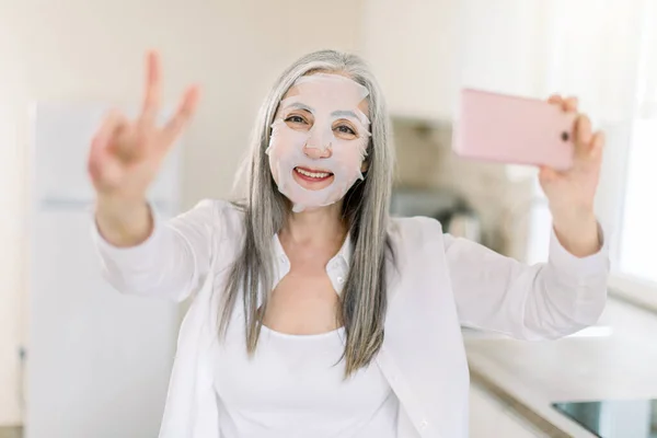 Εσωτερικό πορτρέτο της χαμογελαστής ηλικιωμένης γυναίκας με μάσκα προσώπου και αντιγήρανσης, ποζάρει στην κάμερα στο σπίτι, κρατώντας το smartphone της και δείχνοντας το σήμα της νίκης. Έννοια ομορφιάς και περιποίησης προσώπου — Φωτογραφία Αρχείου