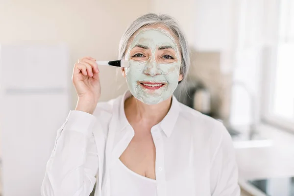 Mulher caucasiana muito idosa com cabelos grisalhos, posando para câmera na moderna cozinha doméstica enquanto aplica máscara facial de barro no rosto com escova cosmética. Spa e beleza, cuidado antienvelhecimento em casa — Fotografia de Stock