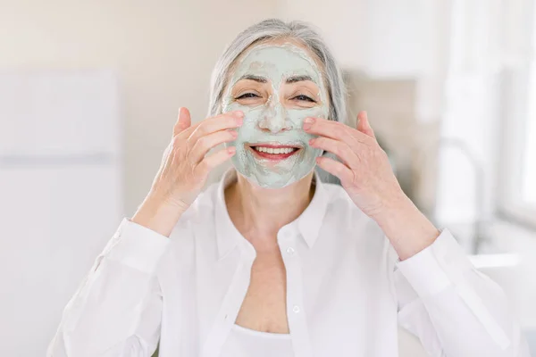 Close up beauty portrait of smiling pretty senior gray haired woman, applying fruit mud nourishing facial mask on her face with hands, posing to camera on the background of home interior — Stock Photo, Image