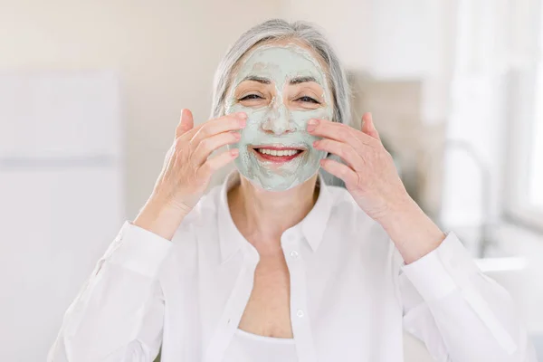 Foto kecantikan wanita berambut abu-abu senior yang tersenyum, mengenakan masker wajah bergizi lumpur buah di wajahnya dengan tangan, berpose ke kamera di latar belakang interior rumah — Stok Foto