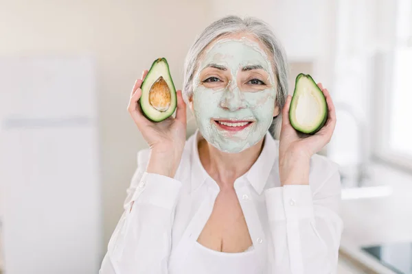 Ομορφιά, αντιρυτιδική και περιποίηση δέρματος έννοια. Κοντινό πλάνο μιας χαρούμενης ηλικιωμένης γυναίκας με άσπρα μαλλιά, με μάσκα προσώπου από πηλό λάσπης, που δείχνει κομμάτια αβοκάντο. Promo μάσκες ανύψωσης προσώπου — Φωτογραφία Αρχείου