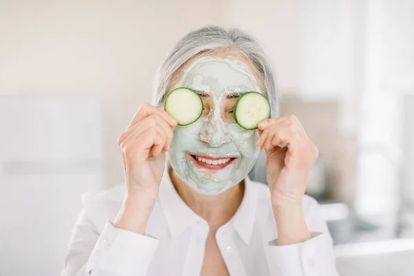 Primer plano de una hermosa mujer jubilada con máscara facial en su cara sosteniendo rebanadas de pepino fresco cubriendo sus ojos, sobre el fondo interior del hogar. Cuidado y tratamiento de la piel, spa en casa — Foto de Stock