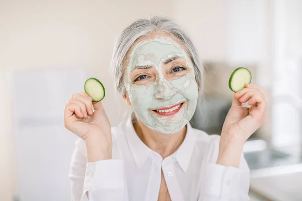 Tubuh dan perawatan kulit wajah, pengobatan anti penuaan. Menutup gambar horisontal wanita cantik dengan masker tanah liat, memegang potongan mentimun, sambil berdiri di atas interior rumah latar belakang — Stok Foto