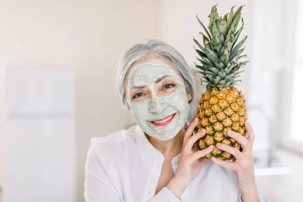 Promoción de hidratante mascarilla anti-envejecimiento nutritiva, espacio de copia para el anuncio. Primer plano retrato de belleza de la mujer sonriente bastante mayor con barro verde máscara facial de barro, la celebración de piña fresca —  Fotos de Stock