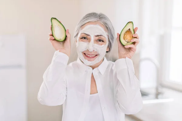 Retrato de mulher idosa atraente sorrindo segurando duas metades de abacate, com máscara de barro facial nutritiva, posando sobre fundo da cozinha casa borrada. Cuidados com a pele e máscaras faciais para antiidade — Fotografia de Stock