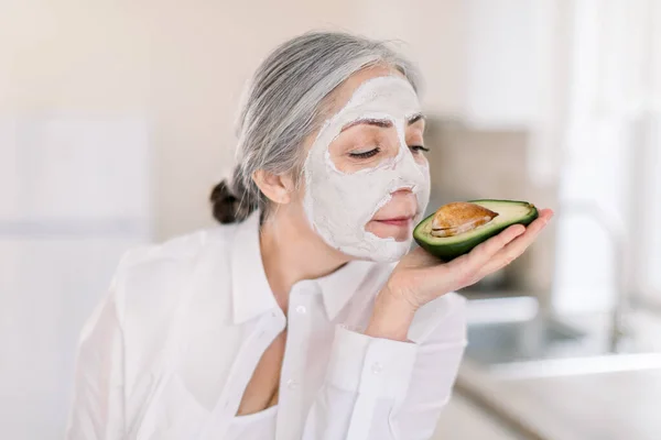 Primer plano de hermosa señora de pelo gris senior con máscara cosmética de arcilla blanca en su cara, disfrutando del tiempo de spa en casa y oliendo aguacate fresco en sus manos. Cuidado de la piel y mascarillas faciales —  Fotos de Stock