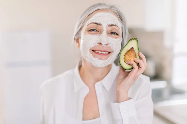 Skin care and anti-aging facial treatment concept. Copy space. Close up beauty portrait of charming senior gray haired woman with white mud facial mask, holding fresh avocado piece, smiling to camera