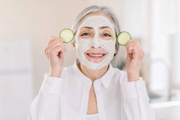 Potret cantik wanita berambut abu-abu senior yang tersenyum dengan masker lumpur wajah dan irisan mentimun di tangannya, berpose dengan latar belakang interior rumah terang. Kosmetik kulit dan konsep spa — Stok Foto