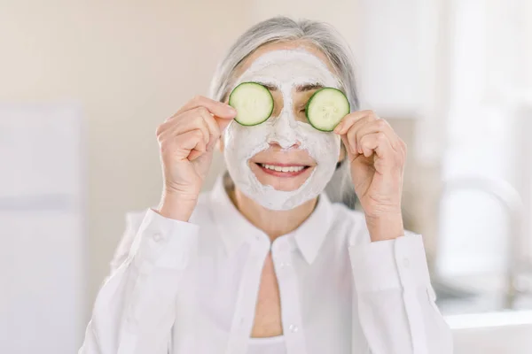 Közelkép egy gyönyörű nyugdíjas nőről, arcmaszkkal az arcán, friss uborkával a kezében, ami eltakarta a szemét, az otthoni belső hátterében. Bőrápolás és öregedésgátló kozmetikai kezelés — Stock Fotó