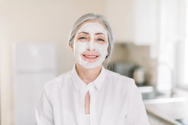 Perawatan kulit dan perawatan kecantikan anti usia, ruang fotokopi. Foto dalam ruangan dari wanita tua berambut pirang cantik dengan tanah liat masker pelembab di wajahnya, berdiri di atas latar belakang dapur ringan di rumah — Stok Foto