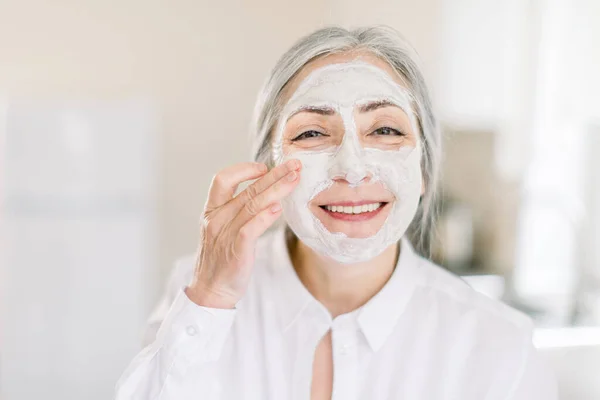 Foto cantik wanita berambut abu-abu senior yang tersenyum, mengenakan masker wajah bergizi lumpur di wajahnya dengan tangan, berpose ke kamera di latar belakang interior rumah — Stok Foto