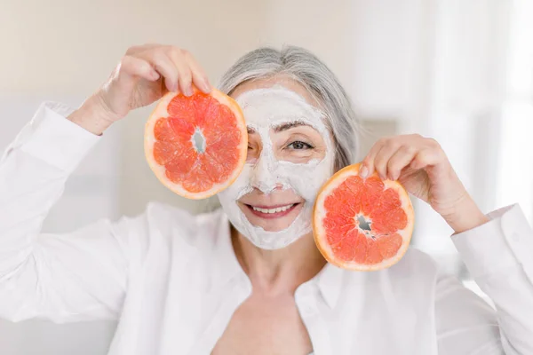 Wanita senior tersenyum gembira dalam kemeja putih, dengan masker tanah liat wajah, berpose ke kamera di interior rumah latar belakang dengan irisan buah anggur segar. Konsep Beauty and Skin Care di Rumah — Stok Foto