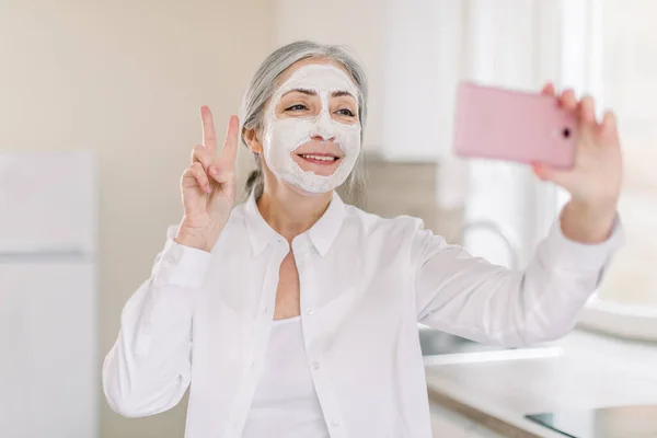 Retrato de senhora sênior positiva, feliz, sorridente com cabelos longos e grisalhos, com máscara facial de lama, se divertindo na cozinha em casa, fazendo foto no celular gesticulando sinal de vitória — Fotografia de Stock