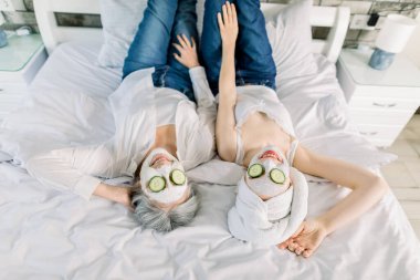 İki çekici kadının üst açılı görüntüsü, yaşlı gri saçlı bayan ve kafasında havlu olan genç kız, yatakta uzanıyor ve yüz maskeleri ve salatalık dilimleriyle spa gününün tadını çıkarıyor.