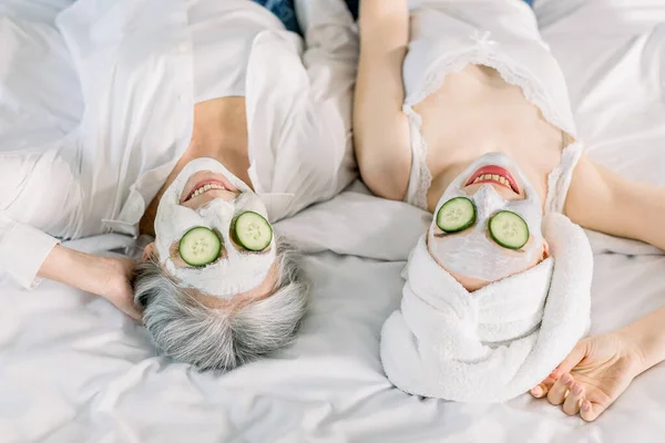 Close up top obraz kobiet, seniora siwe włosy pani matka i jej młoda ładna córka bawiąc się w sypialni i podejmowania gliny maski twarzy z ogórkiem zakrywające oczy. Pielęgnacja skóry — Zdjęcie stockowe