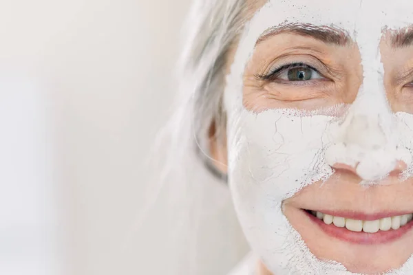 Perawatan kulit dan perawatan kecantikan anti usia. Salin ruang. Wajah wanita berambut abu-abu senior yang cantik dengan masker wajah atau krim di wajahnya, berdiri di atas latar belakang cahaya — Stok Foto