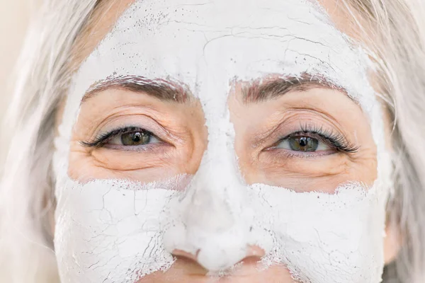 Close up beauty cropped portrait of charming gray haired senior lady, with white facial mask on face, looking at camera while standing on isolated light background. Skin care for elderly women — Stock Photo, Image