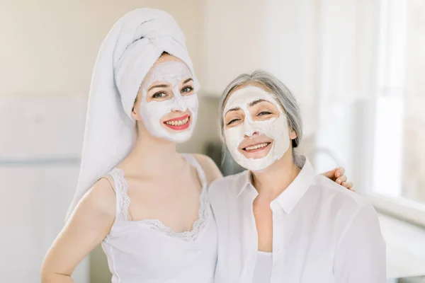 Retrato de duas mulheres caucasianas, menina com cabelo envolto em toalha, e mulher idosa de cabelos grisalhos, com máscaras faciais caseiras em rostos, sorrindo para a câmera, posando em casa. Tratamento da beleza — Fotografia de Stock