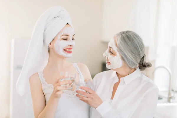 Gaya hidup sehat dan spa keluarga. Gambar horisontal dari dua wanita cantik Kaukasia, nenek dan cucu, memiliki prosedur kecantikan dengan masker tanah liat wajah di rumah dan memegang gelas air — Stok Foto