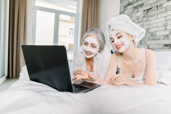 Young attractive woman with towel on head and mud facial mask, lying on bed with her pretty gray haired grandmother, having fun while watching film or talking with friends on laptop