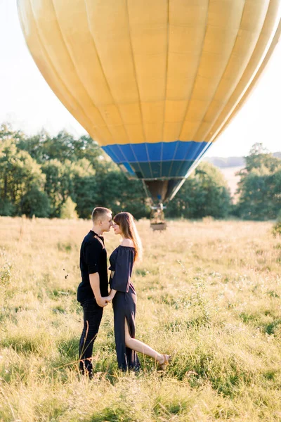 Pareja enamorada se para cara a cara, tomados de la mano, en el campo de verano en el fondo de globo de aire amarillo. Hombre y mujer vestidos de negro, celebrando su aniversario volando en globo — Foto de Stock