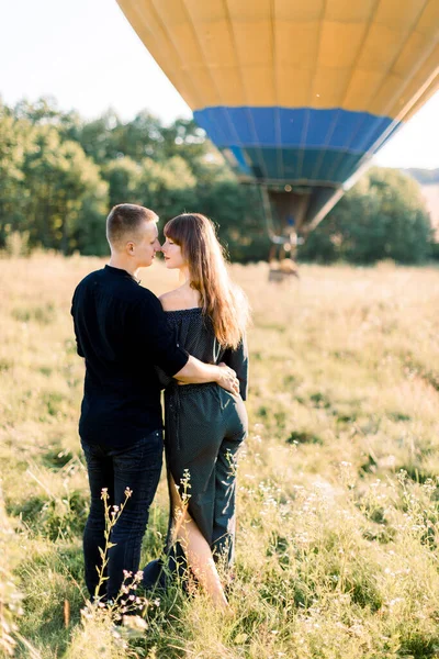 Widok z tyłu pięknej romantycznej pary w czarnych stylowych ubrań, przytulając się, stojąc w lecie słoneczne pole z balonem na ogrzane powietrze na tle. Para jest gotowa na muchę na balonie — Zdjęcie stockowe