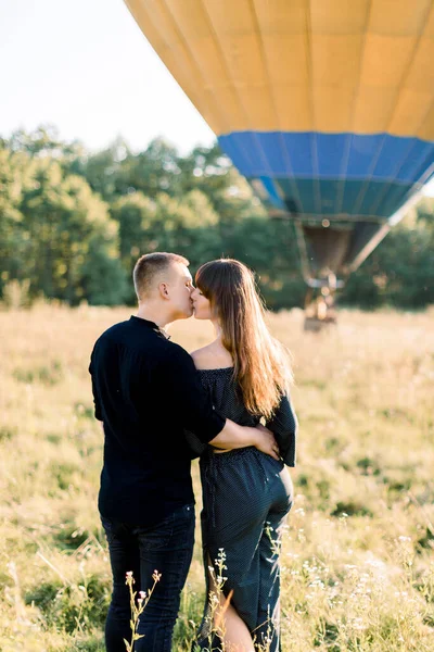 Arka planda, arka planda sıcak hava balonuyla güneşli bir alanda dikilirken siyah şık kıyafetler içinde birbirlerine sarılan güzel romantik çiftin görüntüsü. Çift balonla uçmaya hazır. — Stok fotoğraf