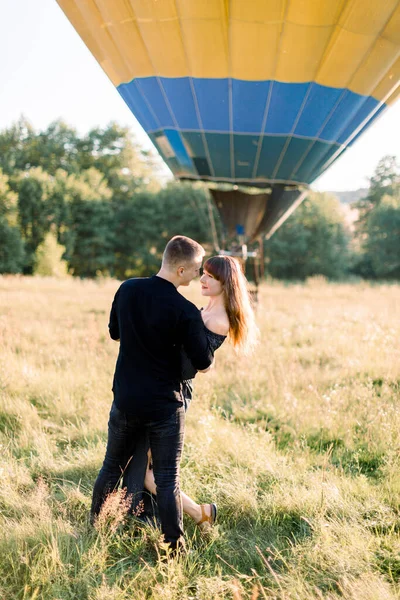 Siyah elbiseli, güzel yeşil çayırlarda sarılıp dans eden, arka planda sıcak sarı hava balonu olan güzel romantik bir çift. Hava balonlu aşk hikayesi, mutlu yıldönümleri. — Stok fotoğraf