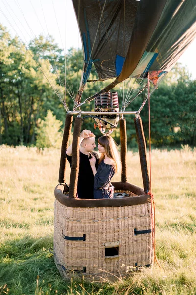 Bonito jovem casal sorridente apaixonado, vestindo roupas casuais pretas, ficando e abraçando em cesta de balão de ar ao pôr do sol, pronto para seu primeiro voo de balão — Fotografia de Stock