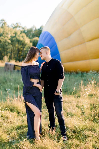 Молодая пара в черной одежде, обнимающая и держащаяся за руки, стоя на летнем поле перед красочным воздушным шаром, готовясь к полету — стоковое фото