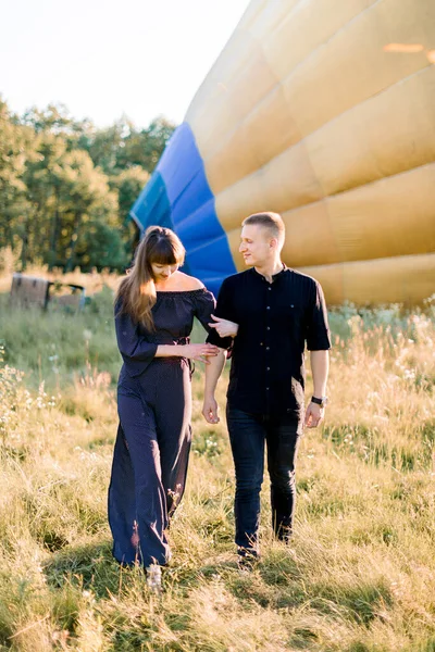 Foto al aire libre de verano de alegre pareja joven caminando durante el atardecer en el campo verde, posando ante la cámara frente al globo de aire caliente amarillo, preparándose para el vuelo — Foto de Stock