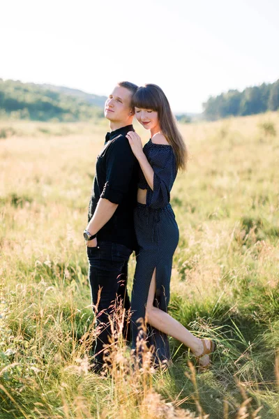 Jeune couple mignon amoureux, portant des vêtements noirs élégants, posant en plein air dans la prairie d'été. Portrait romantique sensuel de jeune couple de mode élégant dans le domaine, femme étreint son homme de dos — Photo