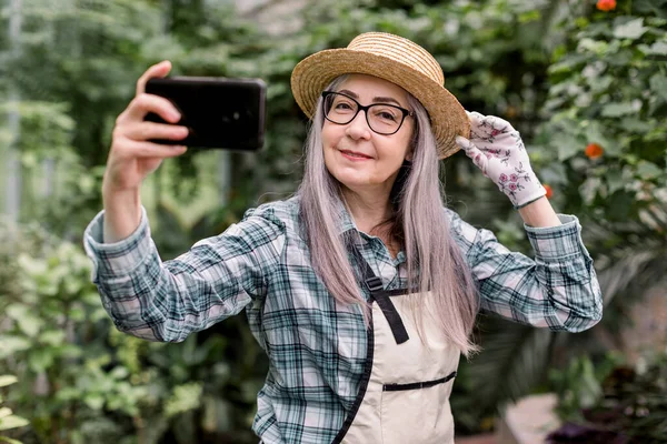 Portrait of attractive cheerful 60s lady gardener with long grey hair, wearing checkered shirt, apron and straw hat, standing in beautiful orangery and having fun while making selfie on smartphone — Stock Photo, Image