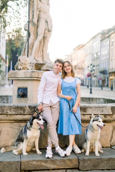 Happy Caucasian jong koppel verliefd, met twee husky honden, poseren voor de camera in het centrum van de oude Europese stad staan in de buurt van oude fontein op stenen trap. Verliefd op honden in de stad — Stockfoto