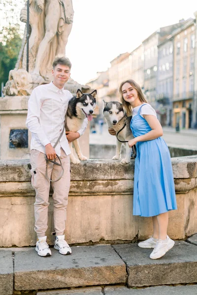 Young stylish couple in casual clothes, posing to camera near ancient stone fountain in the city, hugging their cute husky dogs and smiling. Summer walk in the city with pets — Stock Photo, Image