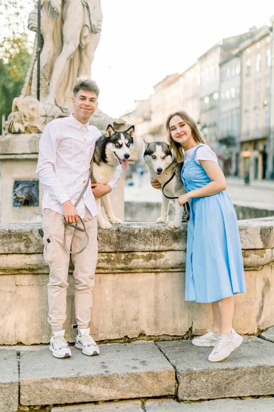 Jeune couple élégant en vêtements décontractés, posant à la caméra près de l'ancienne fontaine de pierre dans la ville, étreignant leurs chiens husky mignons et souriant. Promenade estivale dans la ville avec de beaux chiens — Photo