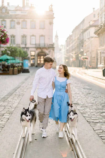 Unga attraktiva par i kärlek, kramar varandra, går på spårvagnsspår med sina två grå husky hundar i staden på en solig sommardag. Gamla byggnader på bakgrunden — Stockfoto