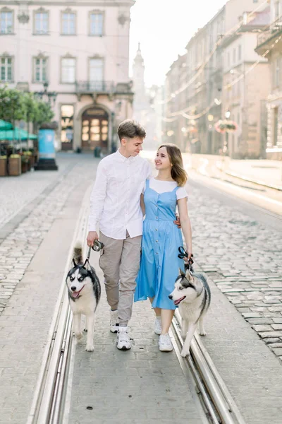 Outdoor shot van jong stel man en vrouw, wandelen met twee grijze husky honden, knuffelen elkaar op de oude Europese stad straat. Buitenactiviteiten, wandelen met dieren concept. — Stockfoto